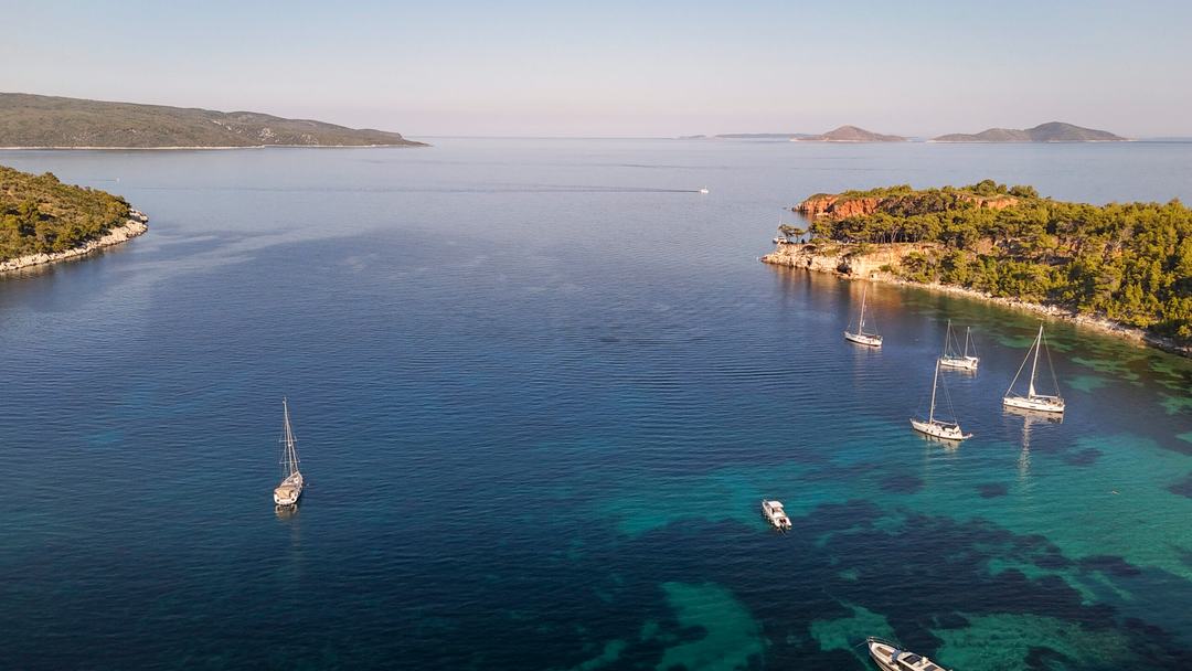 Wide aerial of a calm bay with four sailing yachts anchored off a pine-covered headland with reddish rocks, small motorboats nearby
