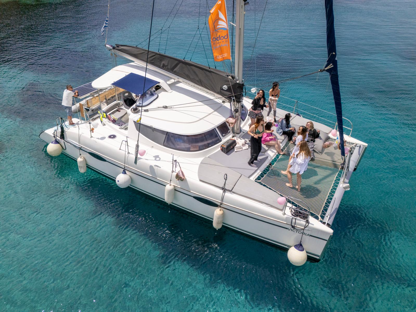 Bachelorette party Halkidiki: aerial of a white catamaran at anchor on turquoise water with a group of women on the foredeck and trampoline, pink and white balloons tied to the rails