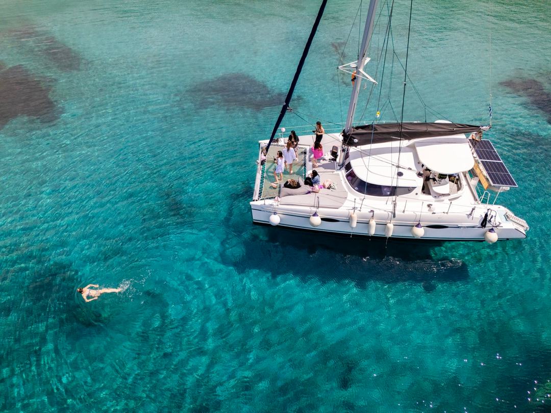Aerial of the white catamaran anchored over turquoise water with guests gathered on the foredeck and one swimmer in the foreground
