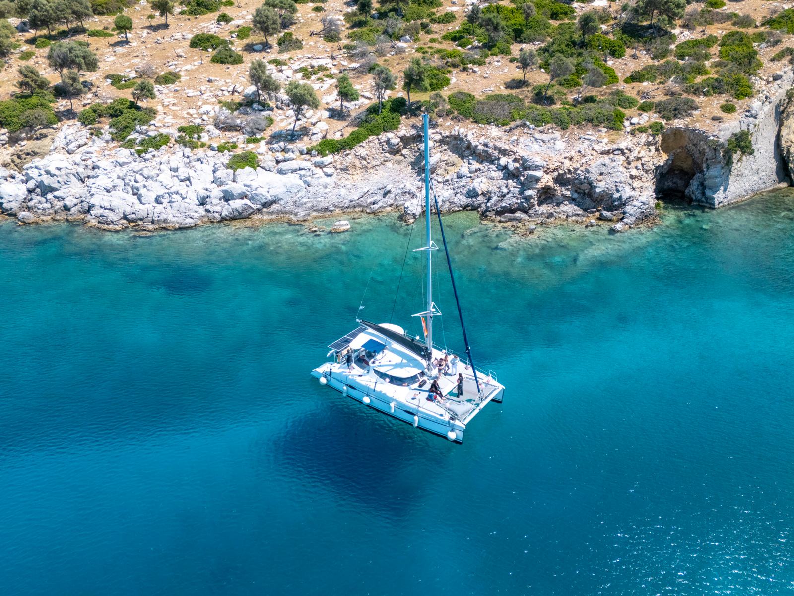 Catamaran full day cruise Halkidiki: aerial of a white catamaran anchored in bright turquoise water beside a pale rocky shoreline with a sea cave, scrub and olive trees