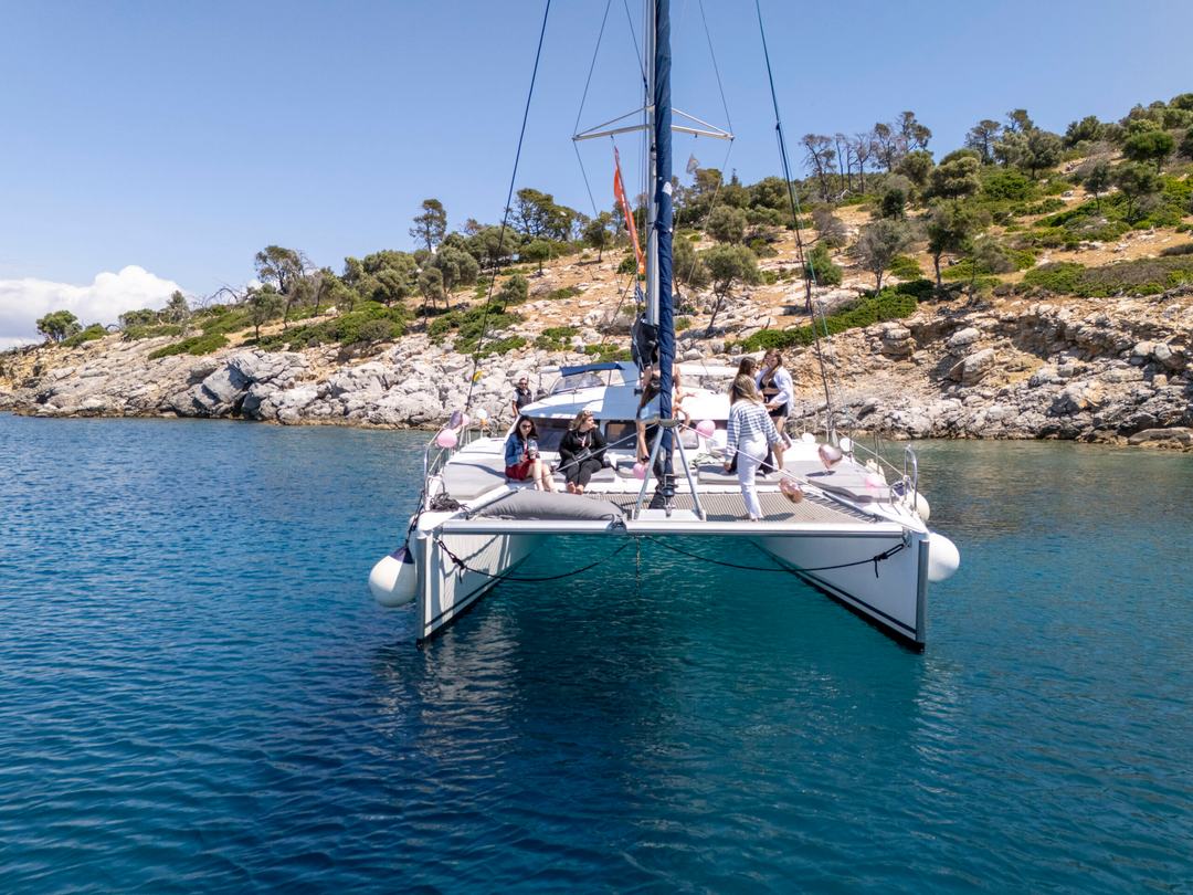 Bow-on view of a white catamaran anchored close to a pale rocky shoreline with olive and pine trees, a group of guests standing on the foredeck and balloons on the rail