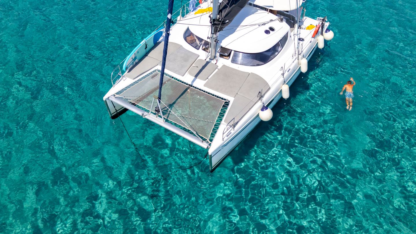 Close aerial of a white catamaran bow with trampoline nets and fenders over crystal-clear turquoise water, one man swimming alongside on a catamaran Halkidiki cruise
