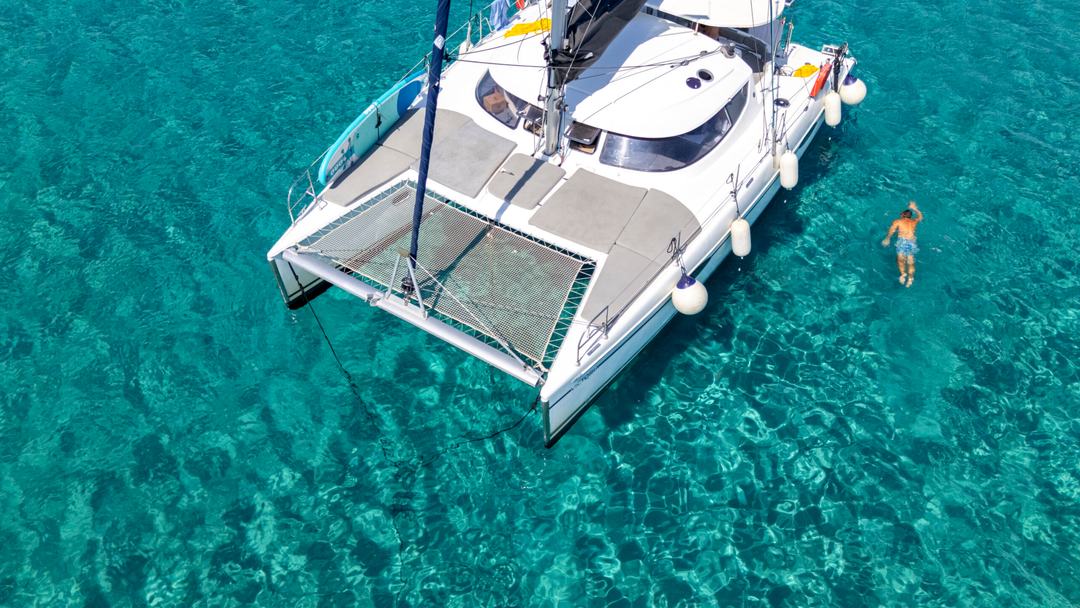 Close aerial of a white catamaran bow with trampoline nets and fenders over crystal-clear turquoise water, one man swimming alongside on a catamaran half day cruise Halkidiki