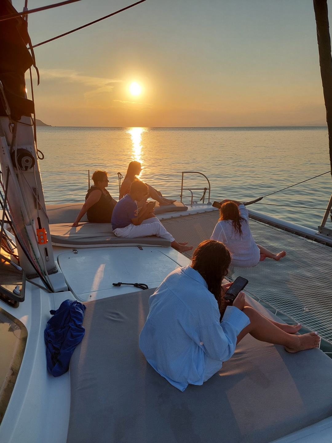 Guests sitting on a catamaran foredeck and trampoline net at sunset, watching the sun low over a calm sea