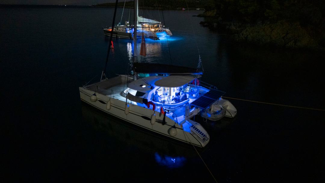 Catamaran at anchor after dark lit by blue cockpit and underwater lights, a second illuminated yacht behind and a dark wooded shore