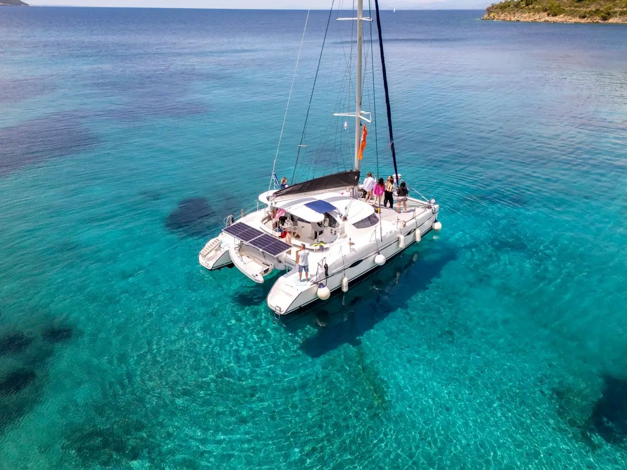 Aerial of a white catamaran with solar panels at anchor over clear turquoise water, guests standing on the foredeck and a low headland in the distance, catamaran half day cruise Halkidiki
