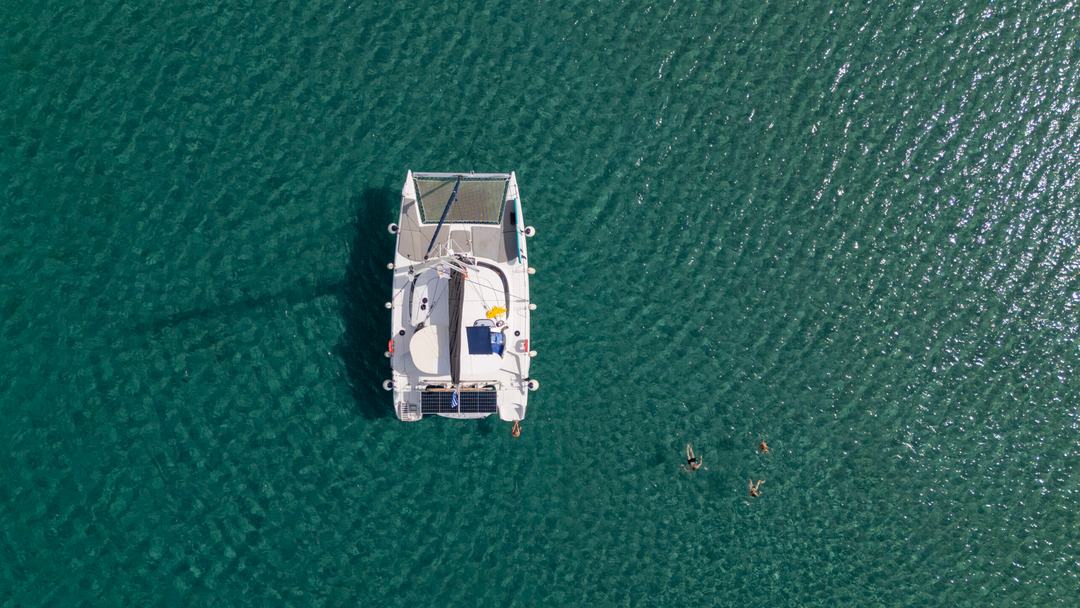 Top-down aerial of a white catamaran anchored over dark green water with three swimmers a short distance away
