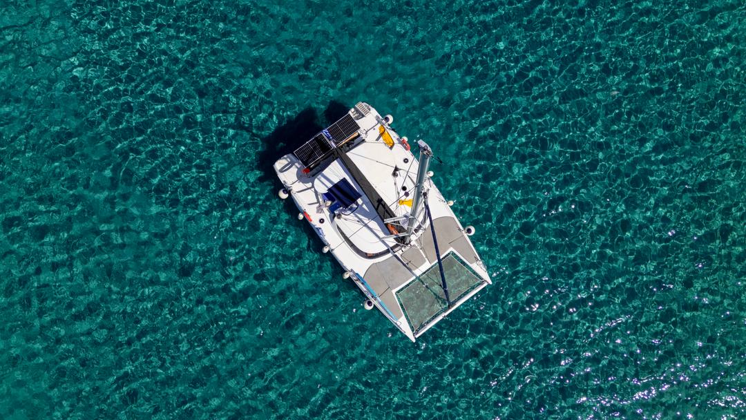 Top-down aerial of a white catamaran with bow trampoline nets anchored over clear green-teal water and a rippled sandy seabed