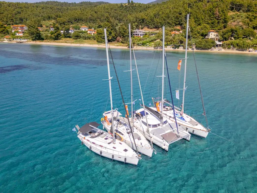 Bachelor party Halkidiki boats: aerial of three sailing yachts and one catamaran rafted side by side at anchor in a calm turquoise bay with a sandy beach and pine-covered hills behind