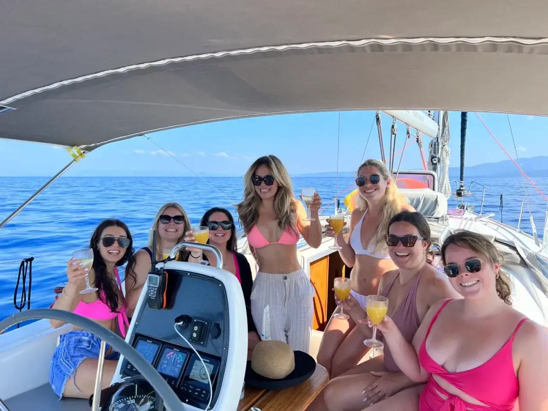 Seven women in swimwear and sunglasses raising cocktail glasses in the cockpit of a sailing yacht under a bimini, open sea behind, on a bachelorette party Halkidiki cruise