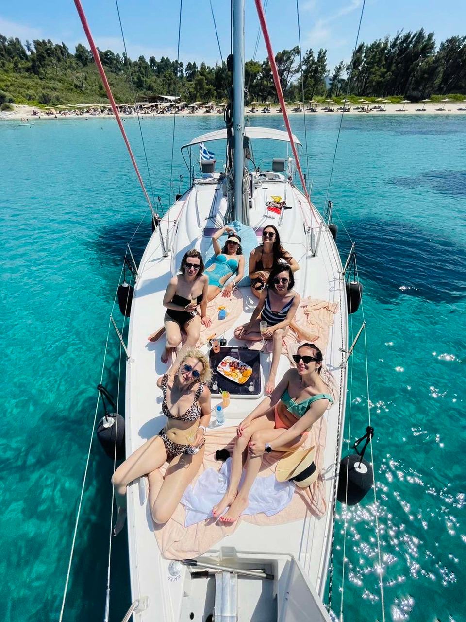 Five women in swimwear and sunglasses posing on the foredeck of a sailing yacht with a snack platter, the yacht anchored over turquoise water off a pine-backed sandy beach on a full day sailing cruise Halkidiki
