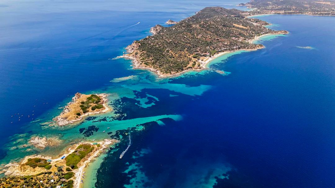 High aerial of a hilly scrub-covered island and smaller rocky islets linked by turquoise shallows, with sandy beaches, deep blue sea and a boat wake in the channel