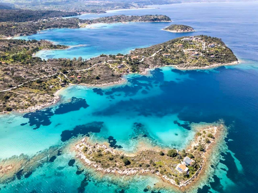 Diaporos Halkidiki: aerial of an indented coastline with small wooded islets, a house on the nearest islet, turquoise lagoon shallows and dark seagrass patches