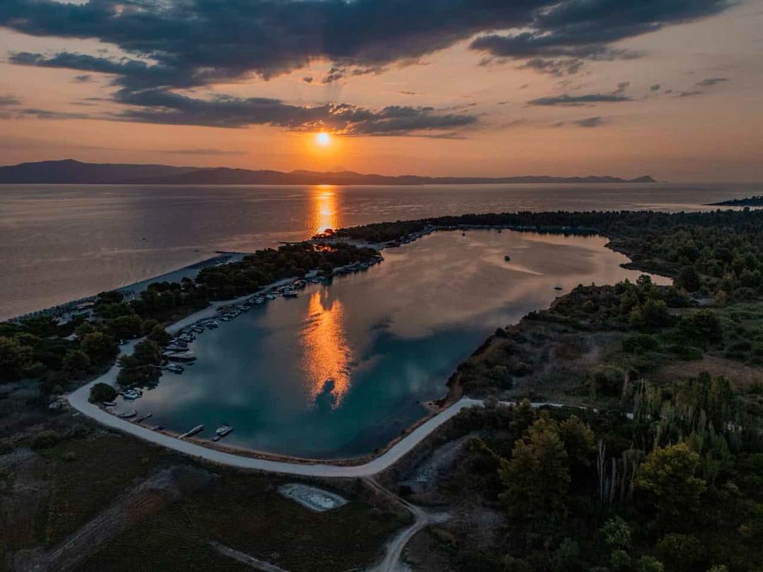 Aerial at sunset over an enclosed lagoon marina with small boats moored along its curved shore, surrounded by pine forest, with open sea and distant mountains behind