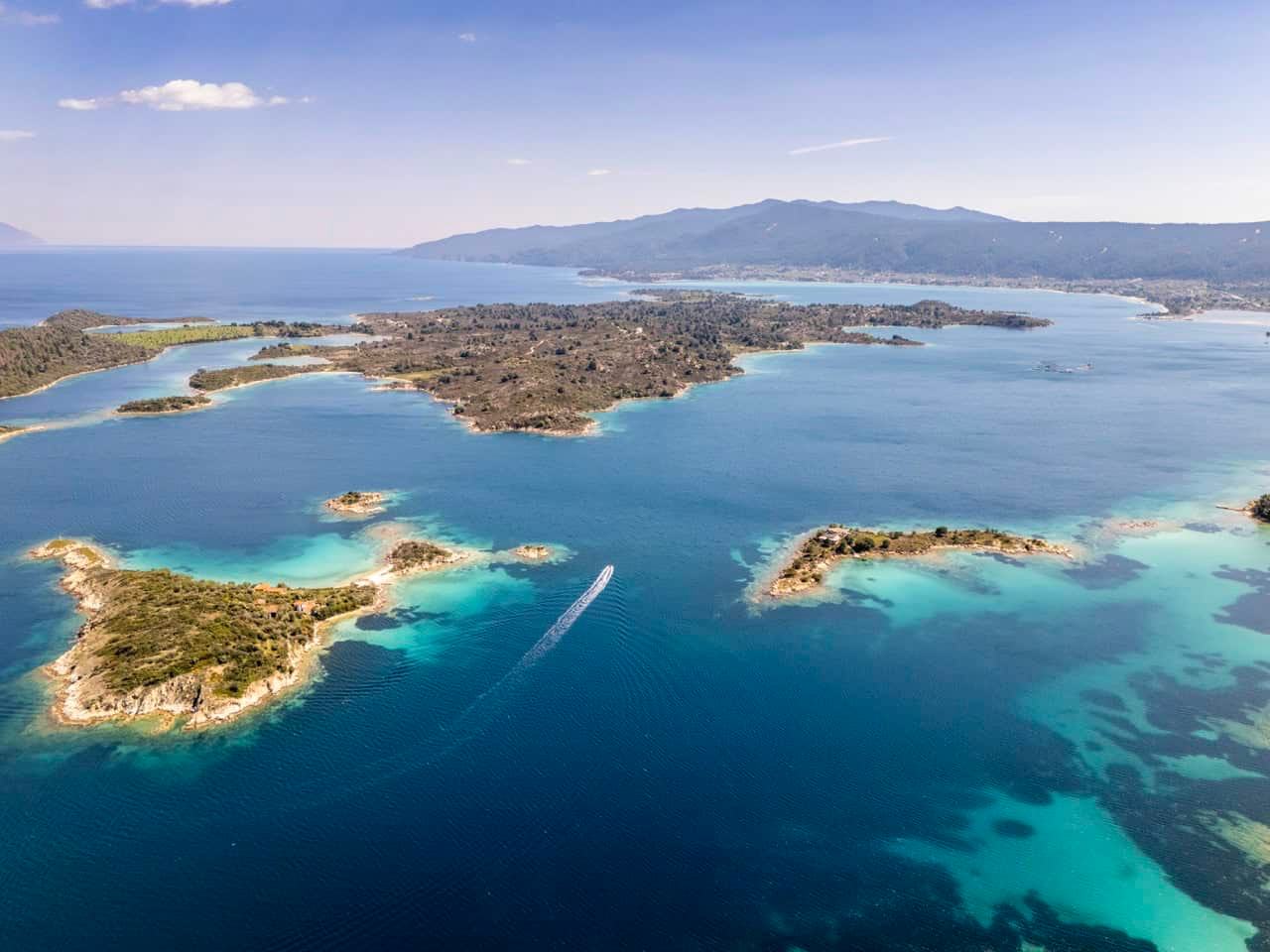 Diaporos Halkidiki: wide aerial of a low pine-covered island and small islets ringed by turquoise shallows, a speedboat wake in the channel and a mountain ridge on the horizon