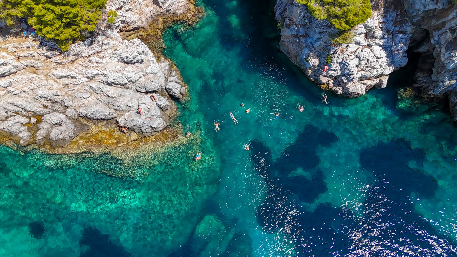 Top-down aerial of a rocky inlet with pale rocks, swimmers in clear turquoise water and a sea cave, a swim stop on a Halkidiki sailing day