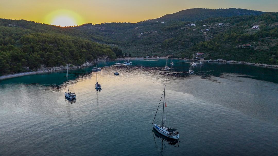 Aerial of a sheltered bay with several sailing yachts at anchor on still water, the sun low over forested hills with scattered houses on the slopes