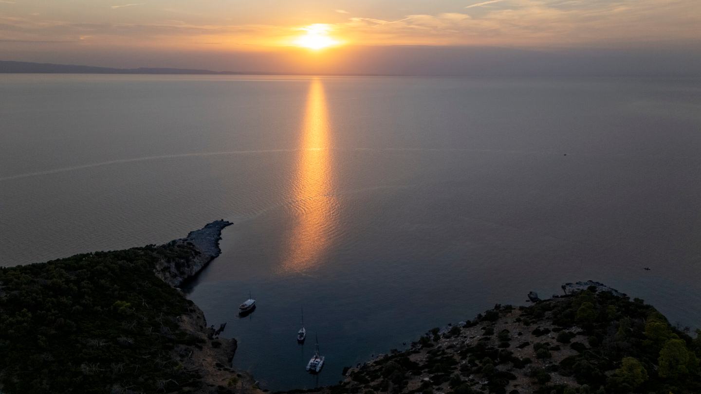 Aerial at sunset of three boats anchored in a small cove between two dark headlands, a long golden reflection across the calm sea