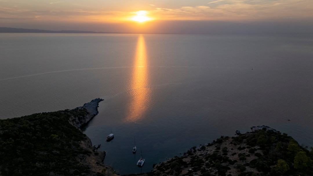 Aerial view at sunset of three boats anchored in a small cove between two dark headlands, a long golden reflection across the calm sea