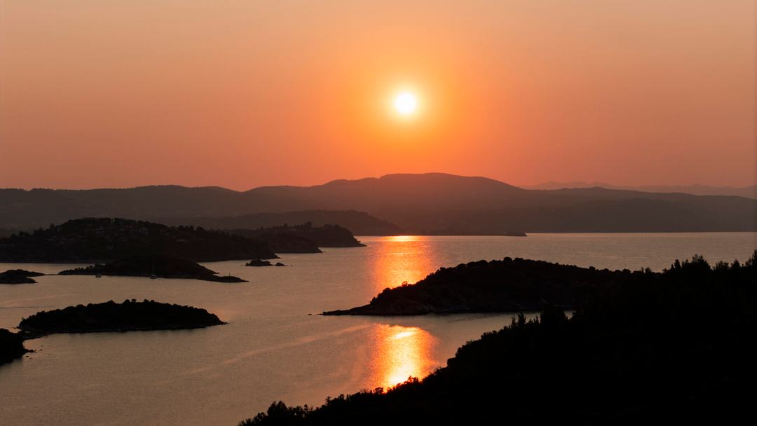 Orange sunset over silhouetted wooded islands and layered hills, the sun mirrored on still water, the light a catamaran sunset cruise Halkidiki is timed for