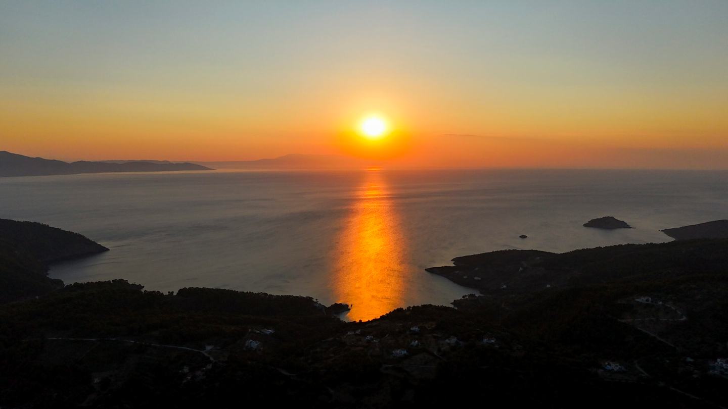 High view over a dark wooded coastline and bay at sunset, the sun low above a hazy sea, the closing light of a catamaran sunset cruise Halkidiki