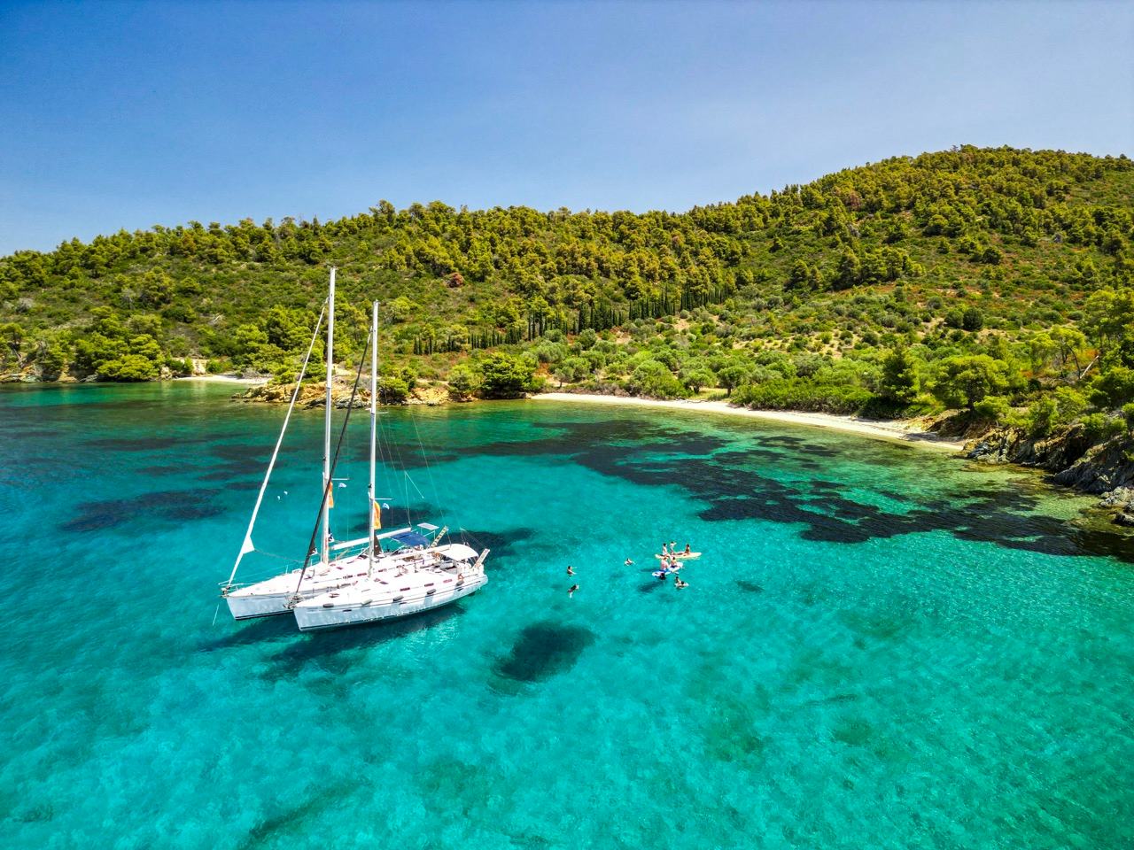 Two white sailing yachts rafted together at anchor in a bright turquoise bay, swimmers and a paddleboard nearby, a sandy beach and pine-covered hillside behind, half day sailing cruise Halkidiki