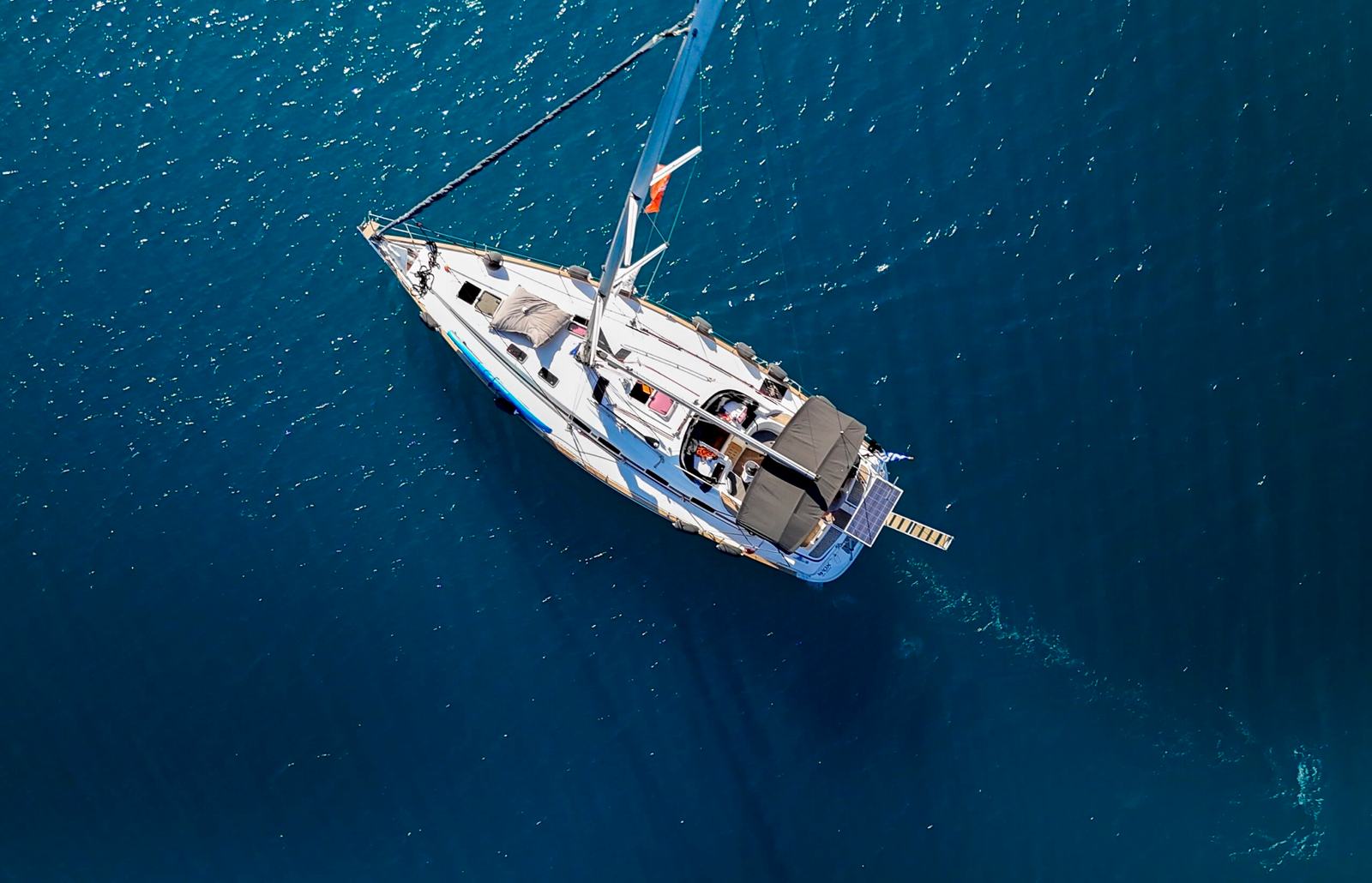 Sailing yacht Halkidiki: aerial view of a white single-hull sailing yacht with a dark bimini on deep blue water, a mooring line running off the bow