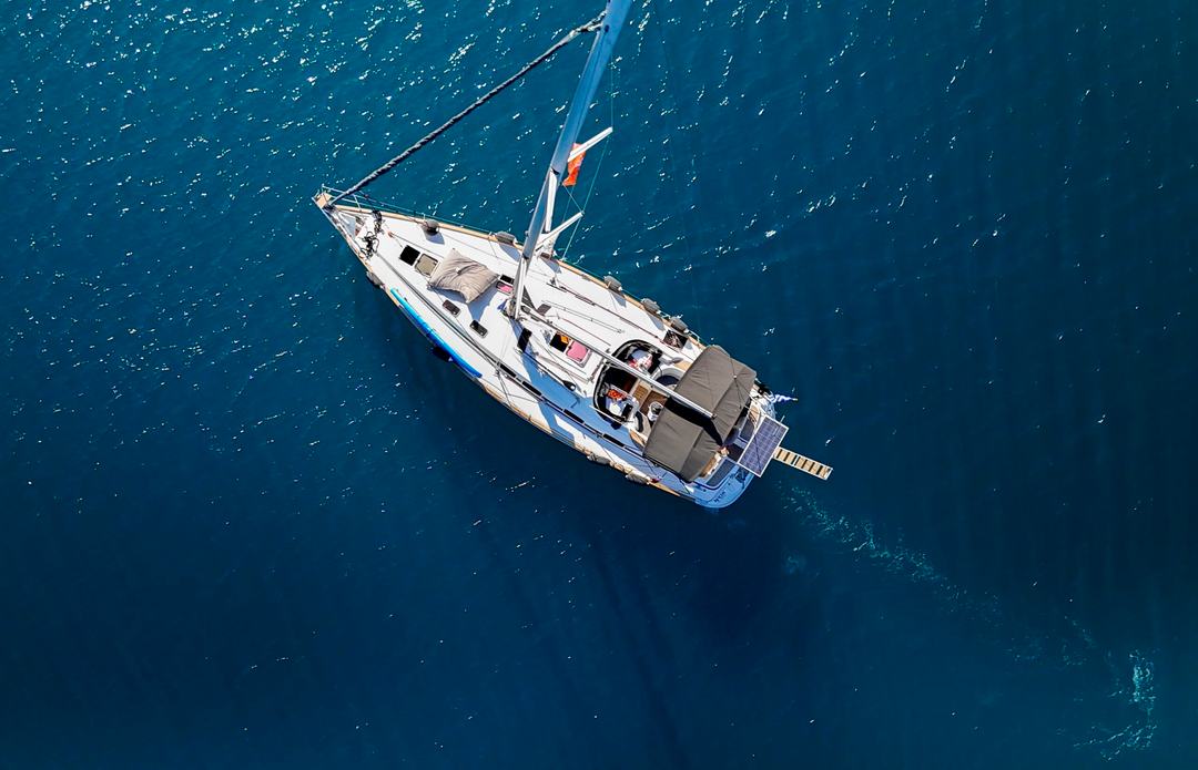 Aerial view of a white single-hull sailing yacht with a dark bimini and a gangway at the stern on deep blue water, a mooring line running off the bow