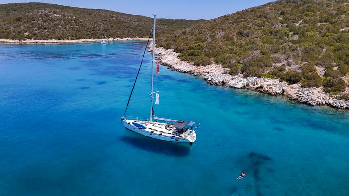 Aerial of a white sailing yacht anchored in a clear blue cove beside a rocky shoreline, full day sailing cruise Halkidiki