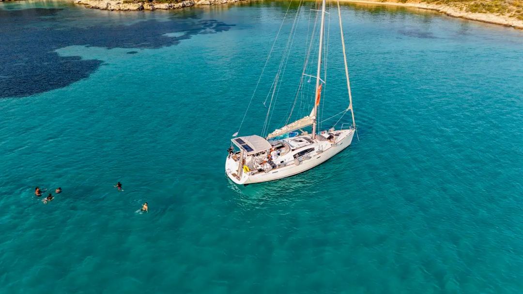 Aerial of a white sailing yacht at anchor in a calm turquoise bay, guests on deck and a group of swimmers and snorkelers nearby, a swim stop on a sailing yacht Halkidiki cruise