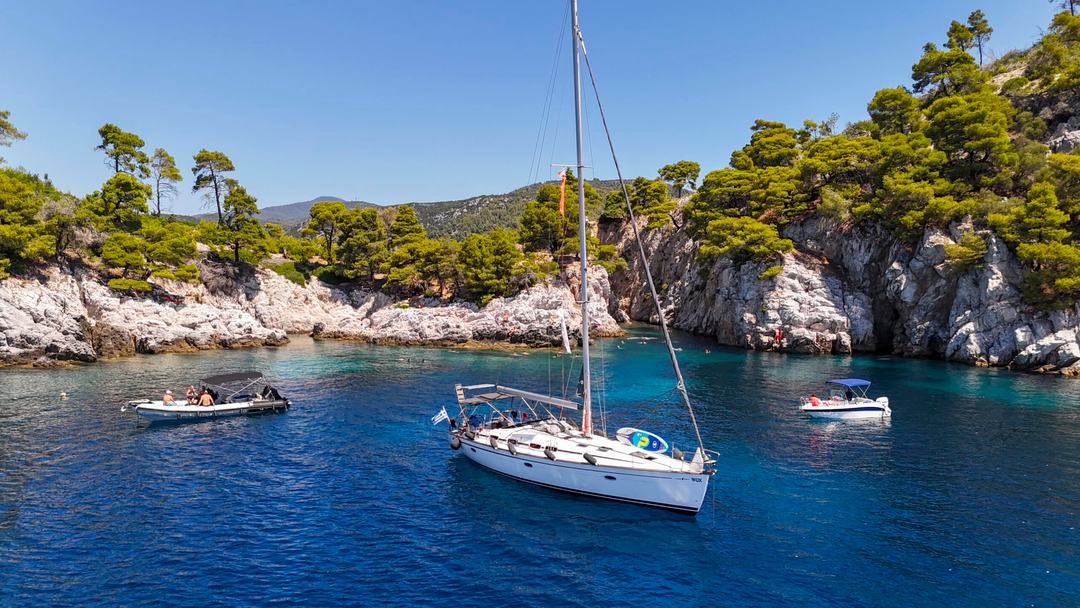 Sailing yacht Halkidiki at anchor: a white sailing yacht in a deep blue rocky cove with pale cliffs topped by pine trees, two small motorboats nearby