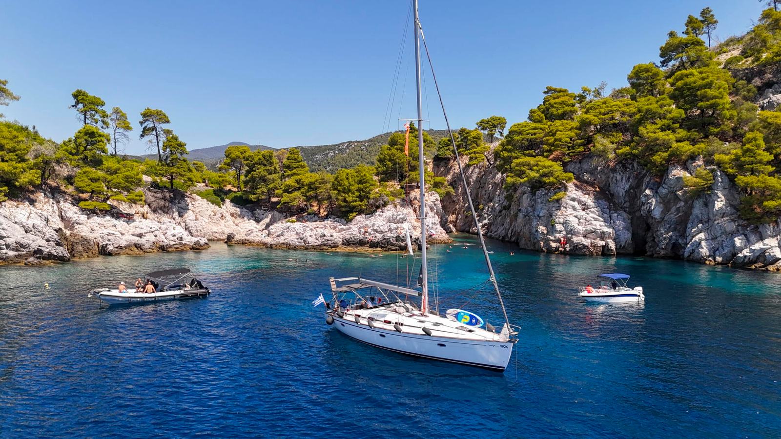 White sailing yacht anchored in a deep blue rocky cove with pale cliffs topped by pine trees, two small motorboats nearby, Halkidiki from Thessaloniki for sailing