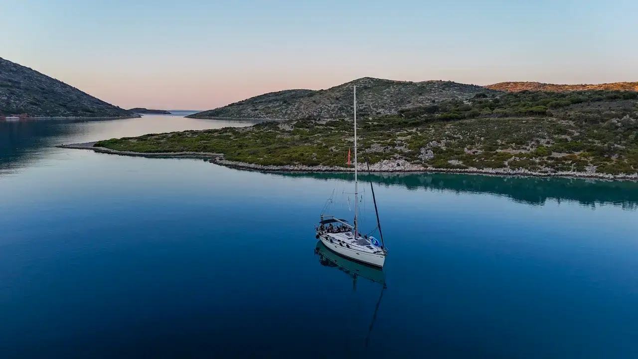 A sailing yacht anchored alone on mirror-calm blue water at dusk, low scrub-covered islands and a pink-tinted sky behind