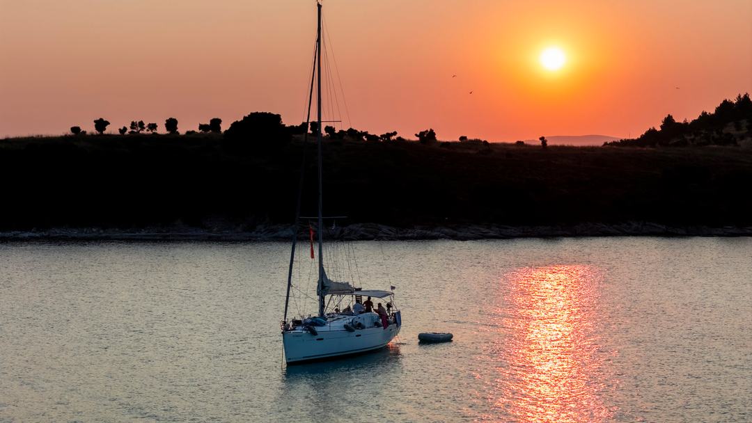 Sailing yacht at anchor with guests in the cockpit and a dinghy astern, a red-orange sun setting over a silhouetted low treeline, the light a sunset sailing cruise Halkidiki is timed for