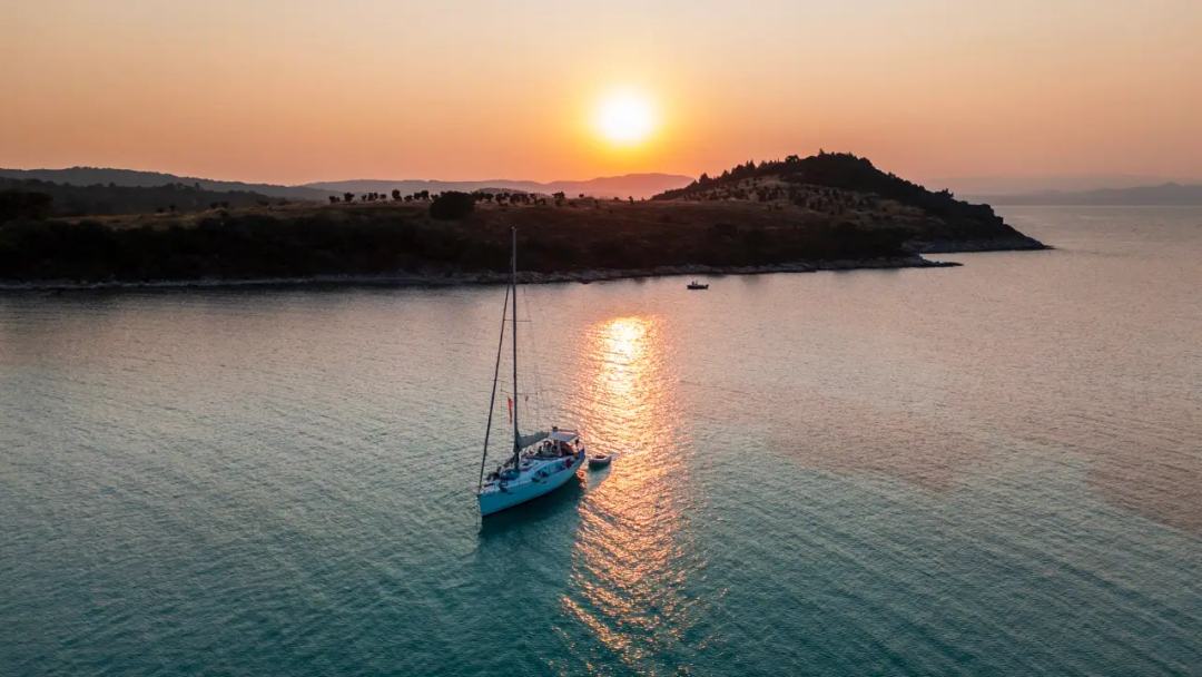 Aerial of a sailing yacht at anchor on calm water at sunset, the sun low over a dark wooded headland with an orange reflection on the sea
