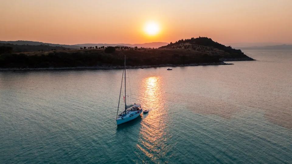 Aerial of a sailing yacht at anchor at sunset off a dark wooded headland, the sun reflected orange on the sea, Halkidiki sailing