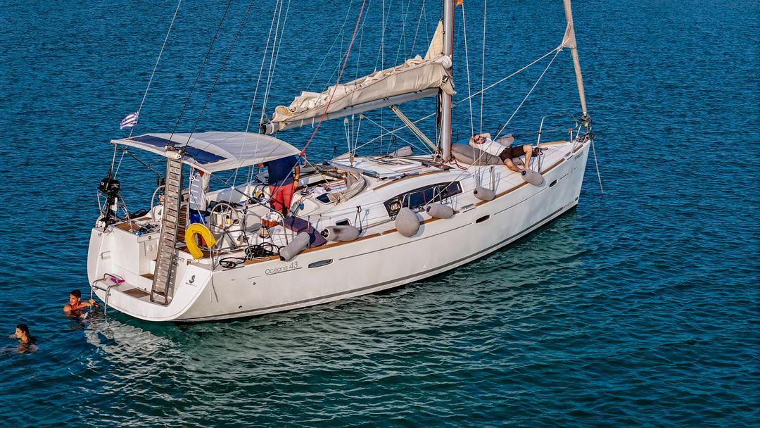 A white single-hull sailing yacht at anchor on blue-green water, swim ladder down with two swimmers at the stern, people on deck and the Greek flag flying