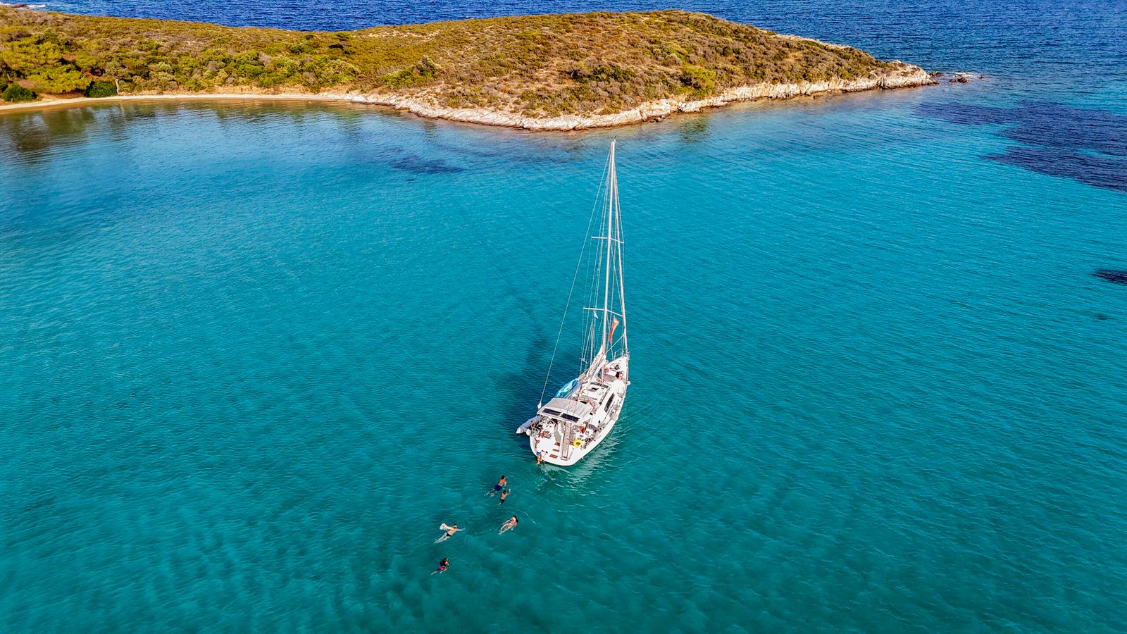 Halkidiki sailing: aerial of a white sailing yacht at anchor in calm turquoise water with swimmers beside it, a low scrub-covered island behind