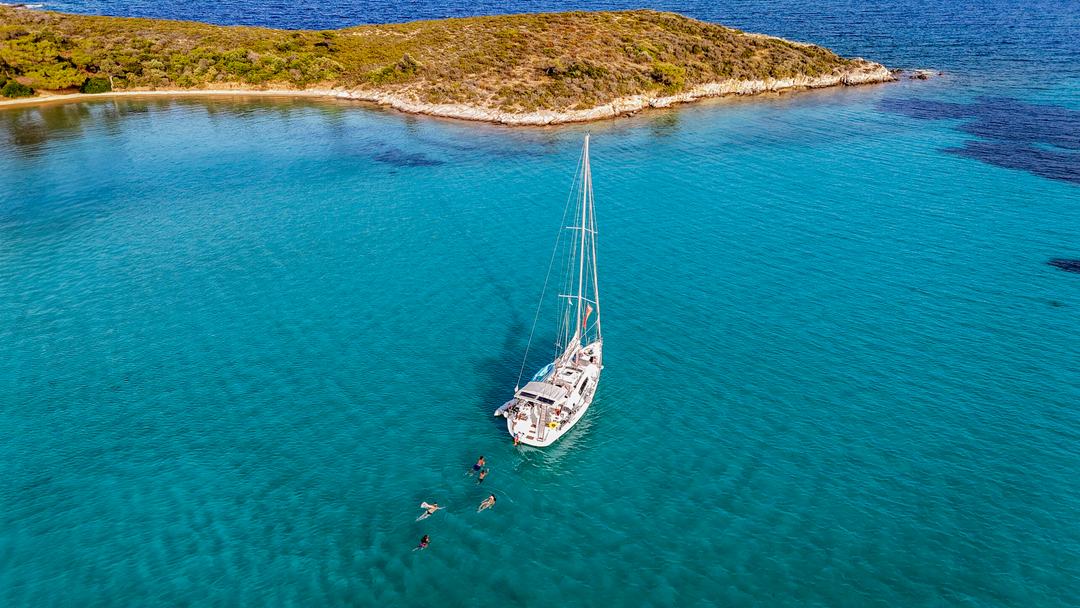 Sailing Diaporos Halkidiki: a white sailing yacht at anchor in calm turquoise water with a few swimmers beside it, a low scrub-covered island with a thin sandy shore behind