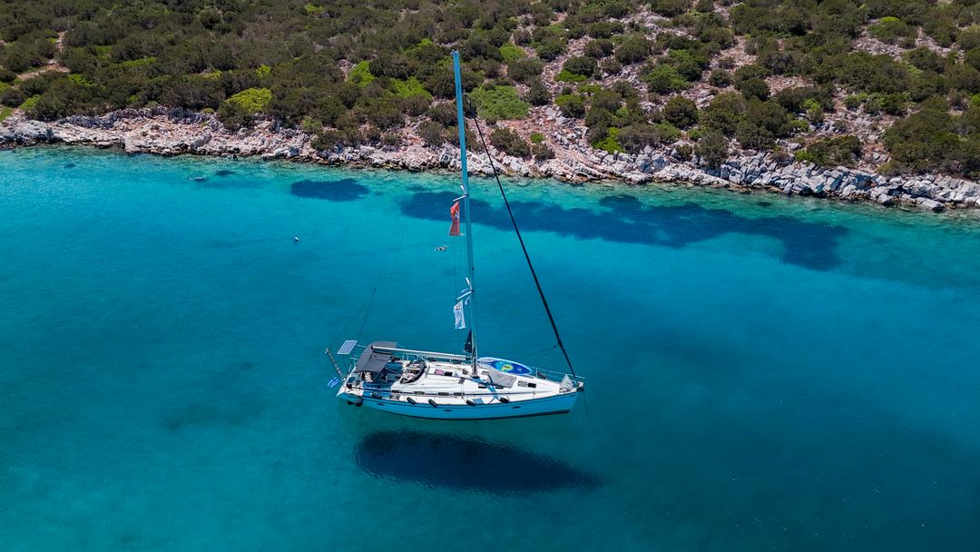 Sailing yacht Halkidiki from above: a white sailing yacht anchored in clear turquoise water just off a rocky, scrub-covered shore