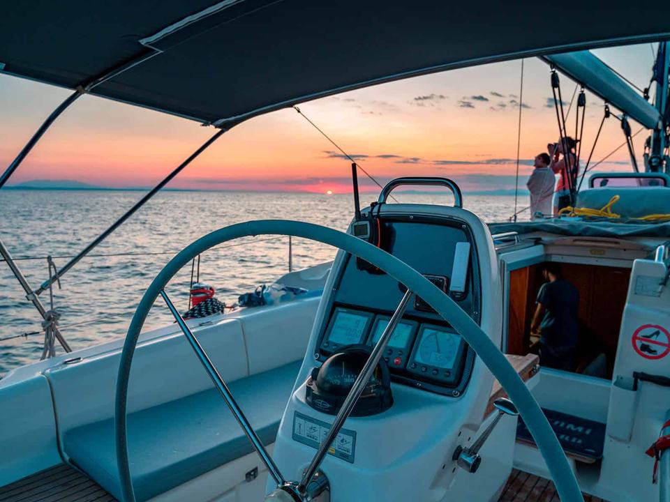 Sailing yacht cockpit with the helm wheel and instrument console in front, a guest photographing a pink sky from the side deck, Halkidiki sailing
