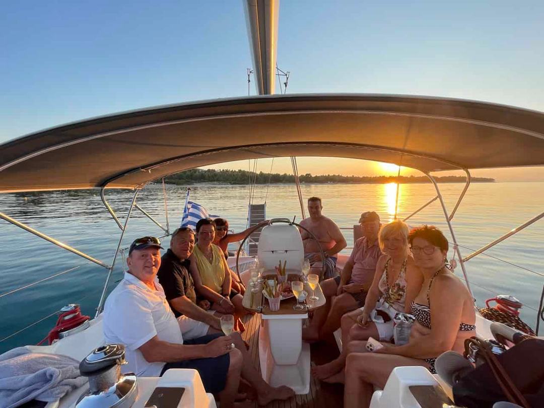 Eight adults seated around the cockpit table of a sailing yacht under a bimini with drinks and snacks on a sunset sailing cruise Halkidiki, the sun setting over a low wooded shoreline behind