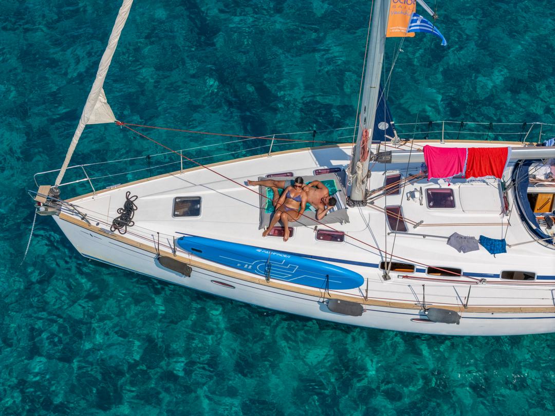 Close aerial of a sailing yacht foredeck over clear green-turquoise water, a blue paddleboard lashed to the rail, towels drying on the lifelines and a Greek flag on the mast