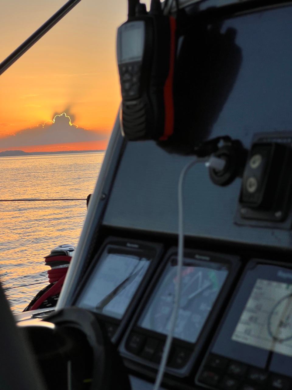 Close-up of a yacht helm console with chartplotter displays and a handheld VHF radio, an orange sky over the sea behind on a sunset sailing cruise Halkidiki