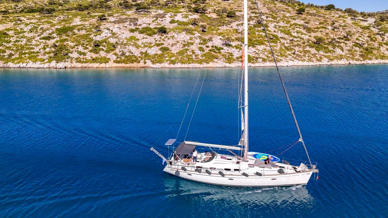 Side-on aerial of a white sailing yacht motoring with sails furled on deep blue water past a barren, scrub-dotted rocky shoreline