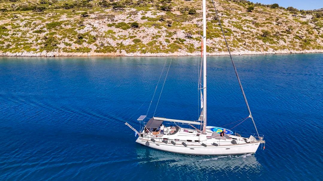 Sailing yacht Halkidiki: side-on aerial of a white sailing yacht motoring with sails furled on deep blue water past a barren, scrub-dotted rocky shoreline