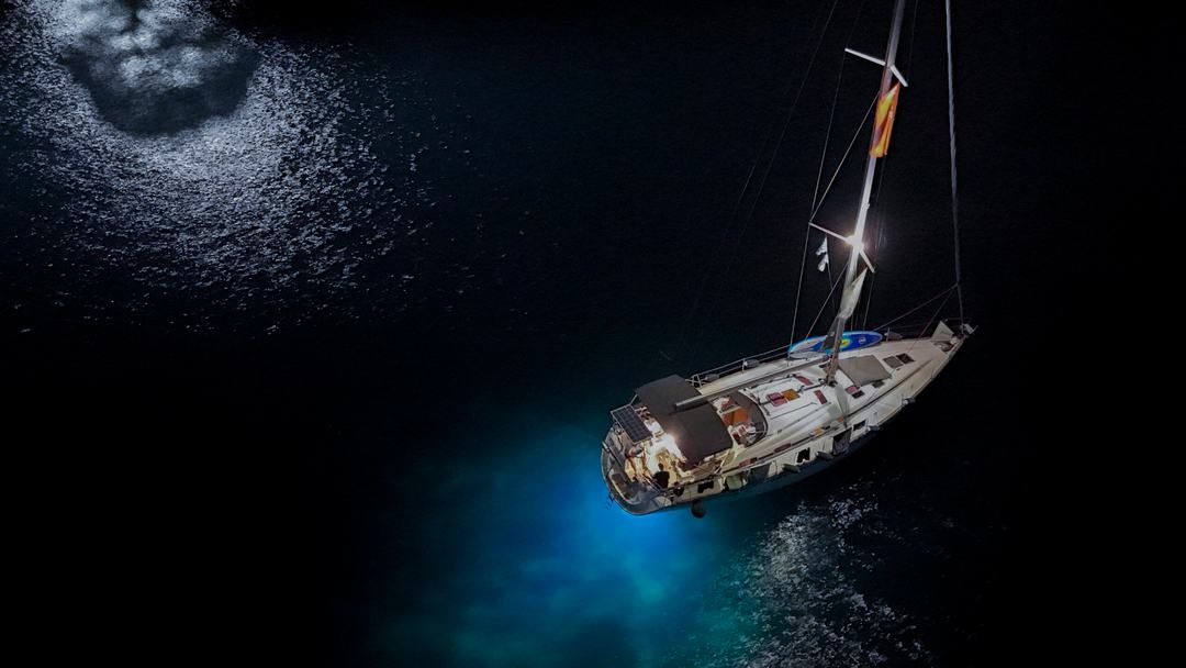 Night aerial of a sailing yacht at anchor with deck lights on and underwater lights casting a blue glow, moonlight reflecting on black water