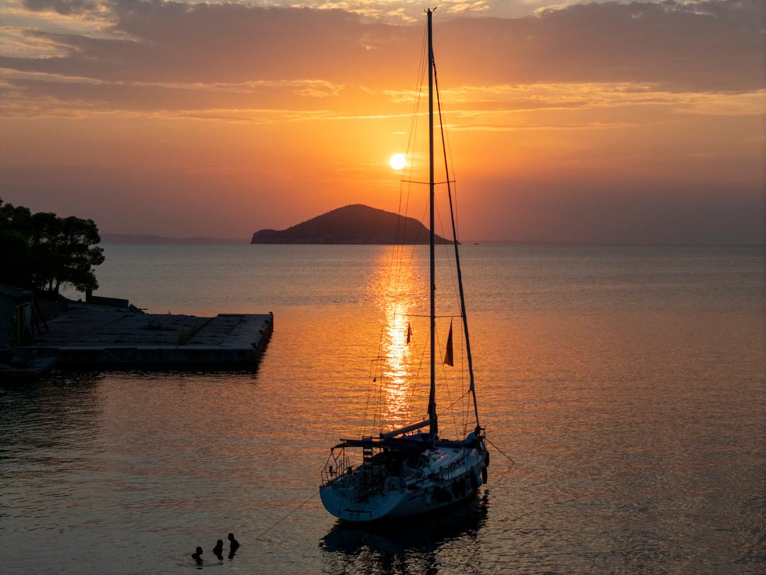Sailing yacht silhouetted at anchor near a concrete pier at sunset, a dome-shaped islet on the horizon under orange clouds and three swimmers in the foreground