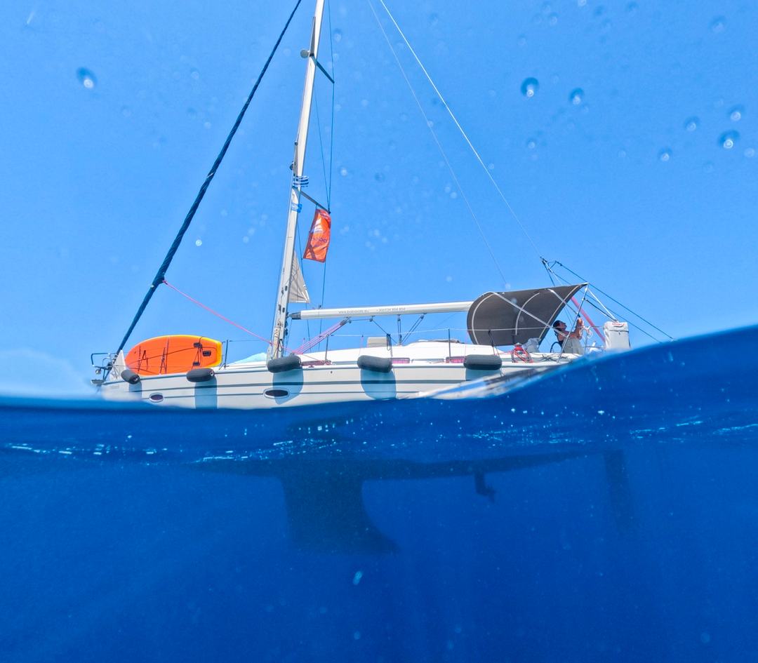 Split over-and-under water shot of a white sailing yacht at anchor, its keel visible beneath a deep blue surface, clear sky above and an orange paddleboard on deck