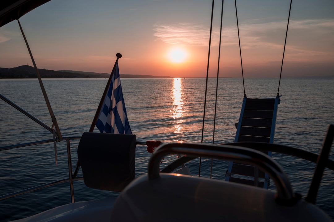 View over the stern of a yacht on a sunset sailing cruise Halkidiki: Greek flag, helm wheel and raised gangway in silhouette, the sun low over a calm sea and a hilly coastline